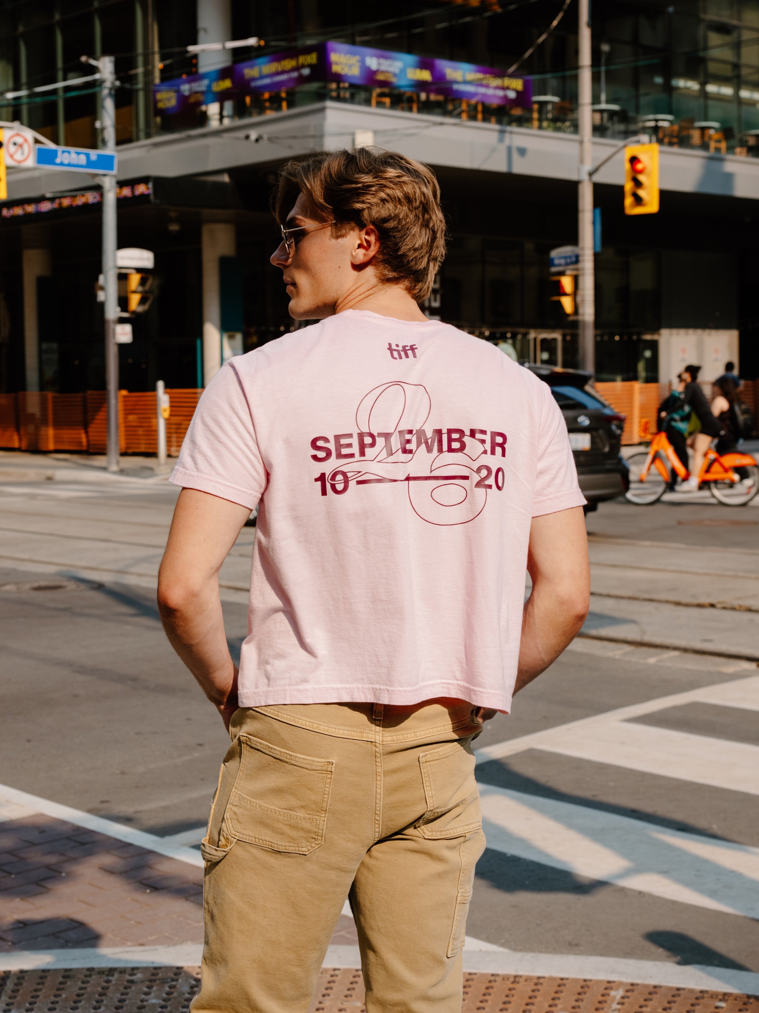 A man standing on a city street corner wearing a light pink graphic t-shirt with 'SEPTEMBER 10 20' printed in maroon on the back, paired with khaki cargo pants.