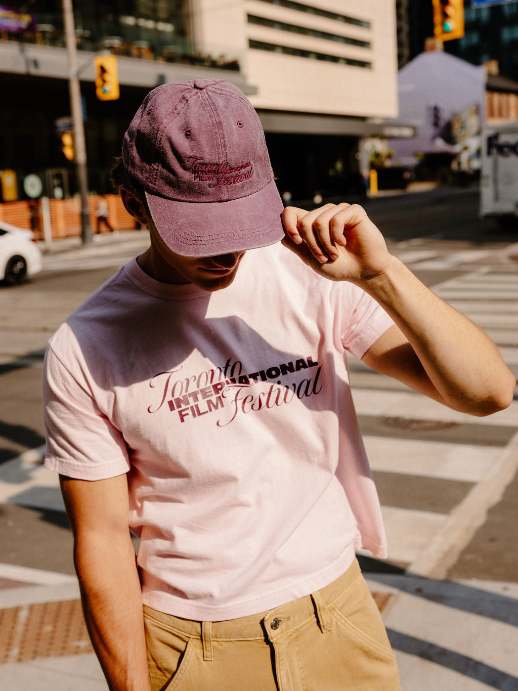 A person standing on a city street wearing a light pink graphic T-shirt with 'Toronto International Film Festival' text, tan pants, and a mauve baseball cap.