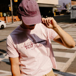 A person standing on a city street wearing a light pink graphic T-shirt with 'Toronto International Film Festival' text, tan pants, and a mauve baseball cap.