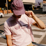 A person standing on a city street wearing a light pink graphic T-shirt with 'Toronto International Film Festival' text, tan pants, and a mauve baseball cap.