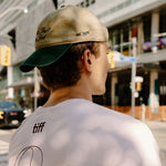A man wearing a white graphic t-shirt and a khaki baseball cap with a green brim, photographed from behind in an outdoor urban setting.