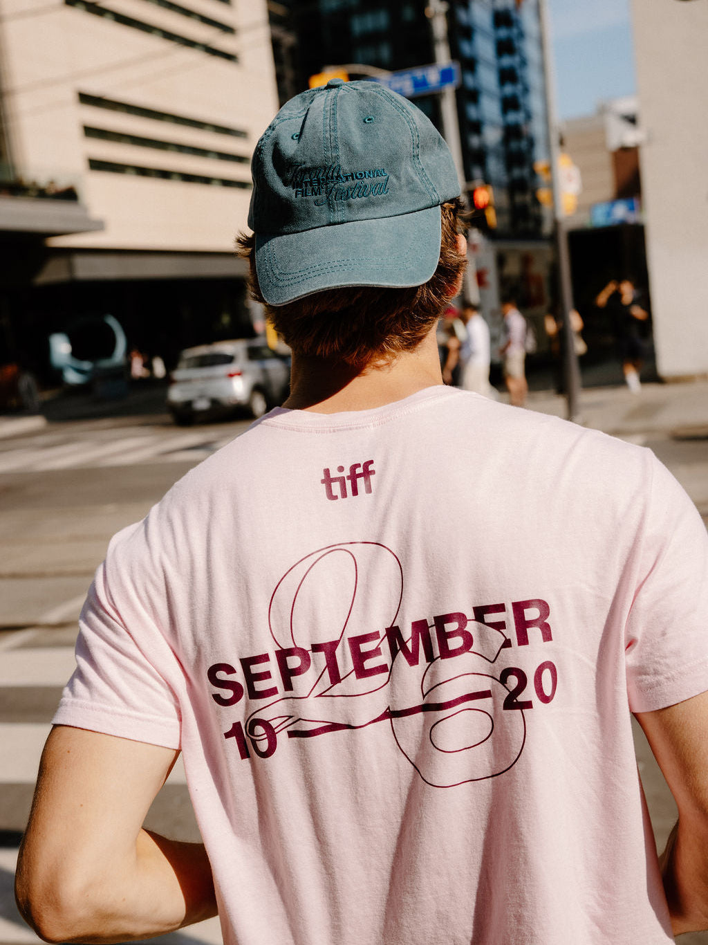 A man viewed from behind wearing a light pink T-shirt with a large dark red graphic on the back reading 'TIFF SEPTEMBER 2020' and a blue baseball cap in an outdoor city street setting.