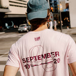 A man viewed from behind wearing a light pink T-shirt with a large dark red graphic on the back reading 'TIFF SEPTEMBER 2020' and a blue baseball cap in an outdoor city street setting.