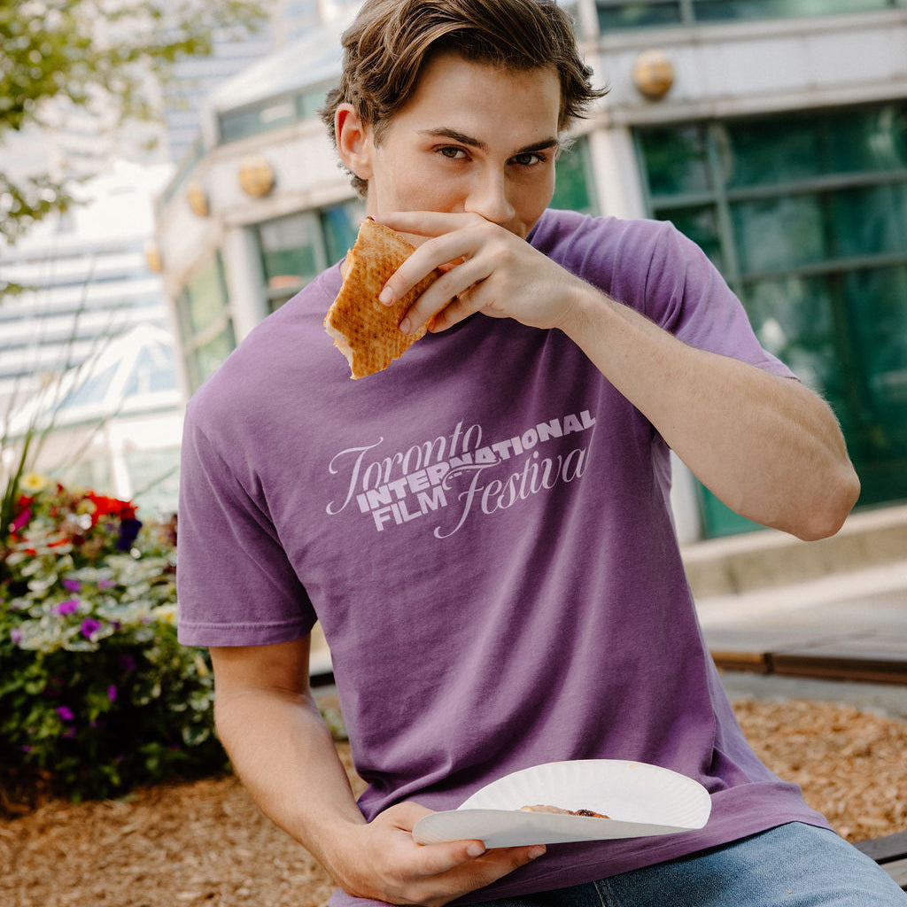 A man wearing a purple t-shirt with 'Toronto International Film Festival' printed in white script, sitting outdoors and eating a sandwich.