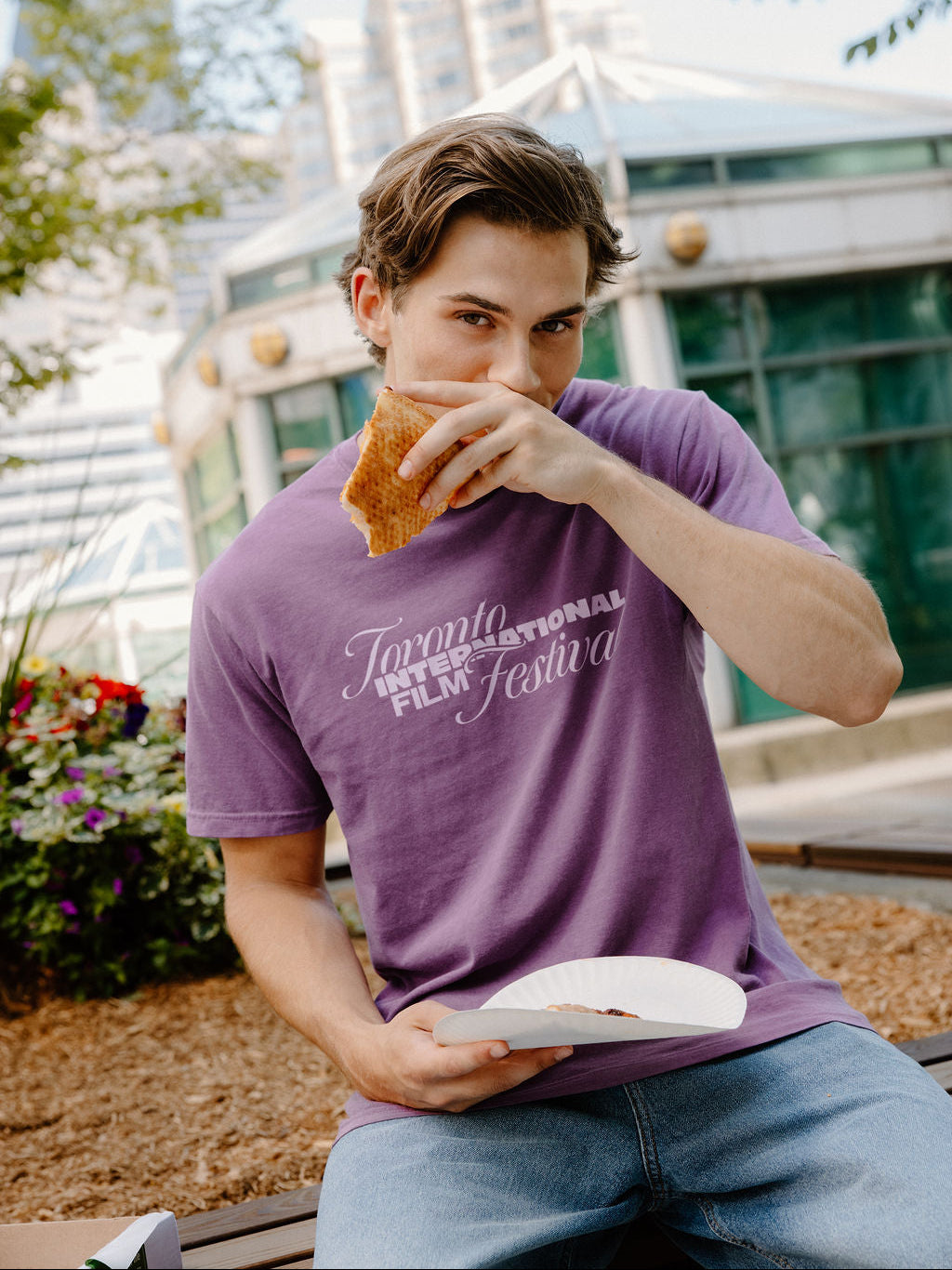 A man wearing a purple t-shirt with 'Toronto International Film Festival' printed in white script, sitting outdoors and eating a sandwich.