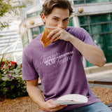 A man wearing a purple t-shirt with 'Toronto International Film Festival' printed in white script, sitting outdoors and eating a sandwich.