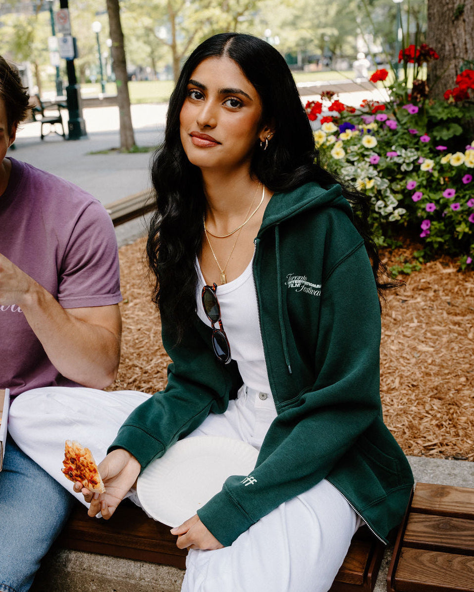 A woman sitting on a wooden bench in an outdoor park setting, wearing a dark green zip-up hoodie over a white top and white sweatpants. A man in a purple t-shirt is partially visible on the left.