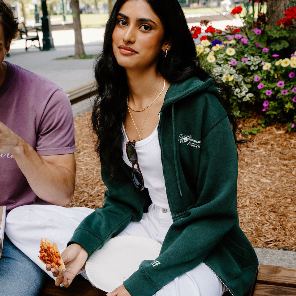 A woman sitting on a wooden bench in an outdoor park setting, wearing a dark green zip-up hoodie over a white top and white sweatpants. A man in a purple t-shirt is partially visible on the left.