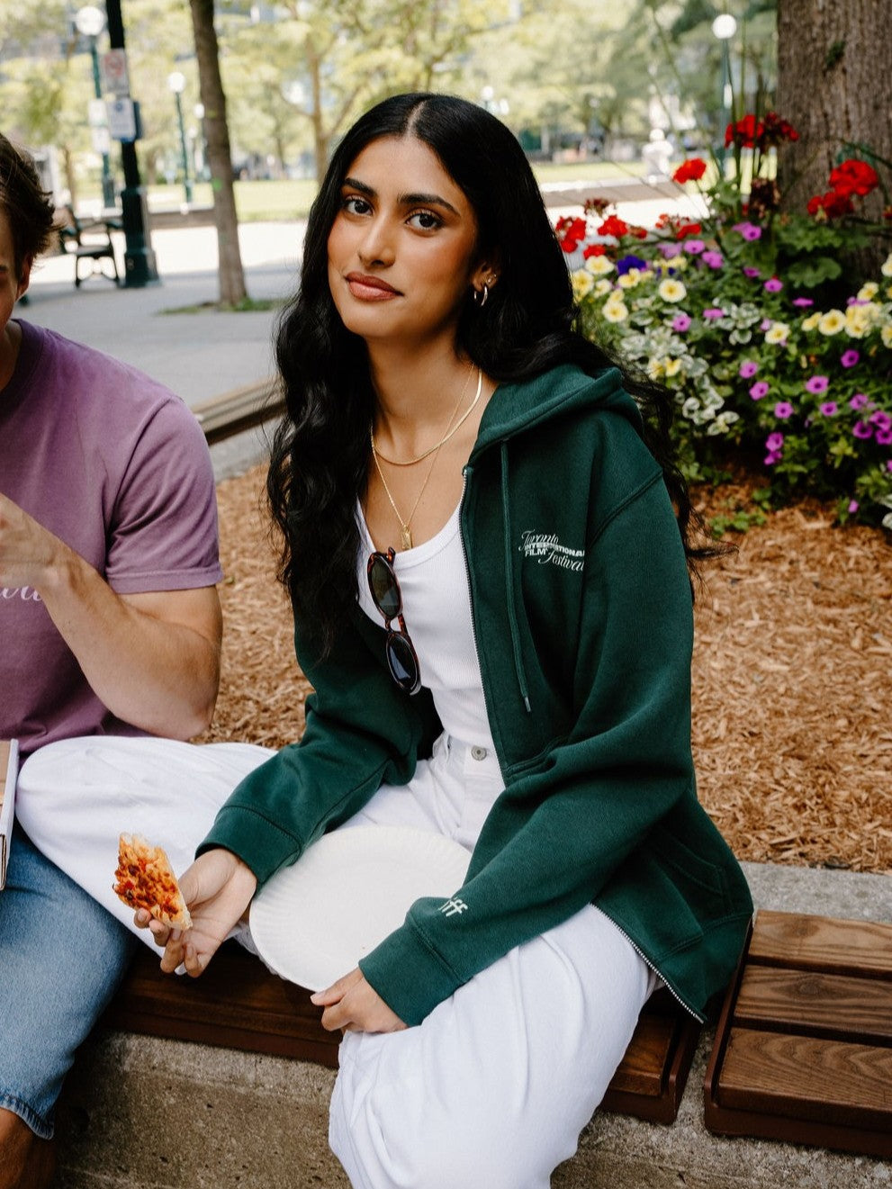 A woman sitting on a wooden bench in an outdoor park setting, wearing a dark green zip-up hoodie over a white top and white sweatpants. A man in a purple t-shirt is partially visible on the left.