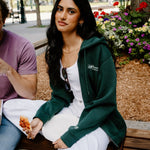 A woman sitting on a wooden bench in an outdoor park setting, wearing a dark green zip-up hoodie over a white top and white sweatpants. A man in a purple t-shirt is partially visible on the left.