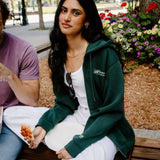 A woman sitting on a wooden bench in an outdoor park setting, wearing a dark green zip-up hoodie over a white top and white sweatpants. A man in a purple t-shirt is partially visible on the left.