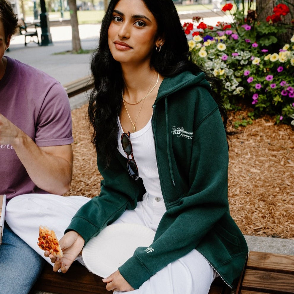 A woman sitting on a wooden bench in an outdoor park setting, wearing a dark green zip-up hoodie over a white top and white sweatpants. A man in a purple t-shirt is partially visible on the left.