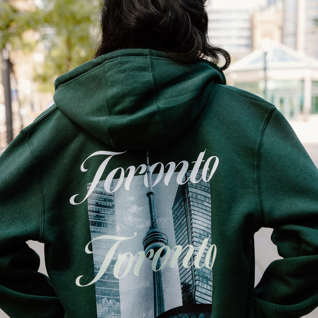 A person viewed from behind wearing a dark green hoodie with a large back graphic featuring the word 'Toronto' repeated in white script over a city skyline and the CN Tower.