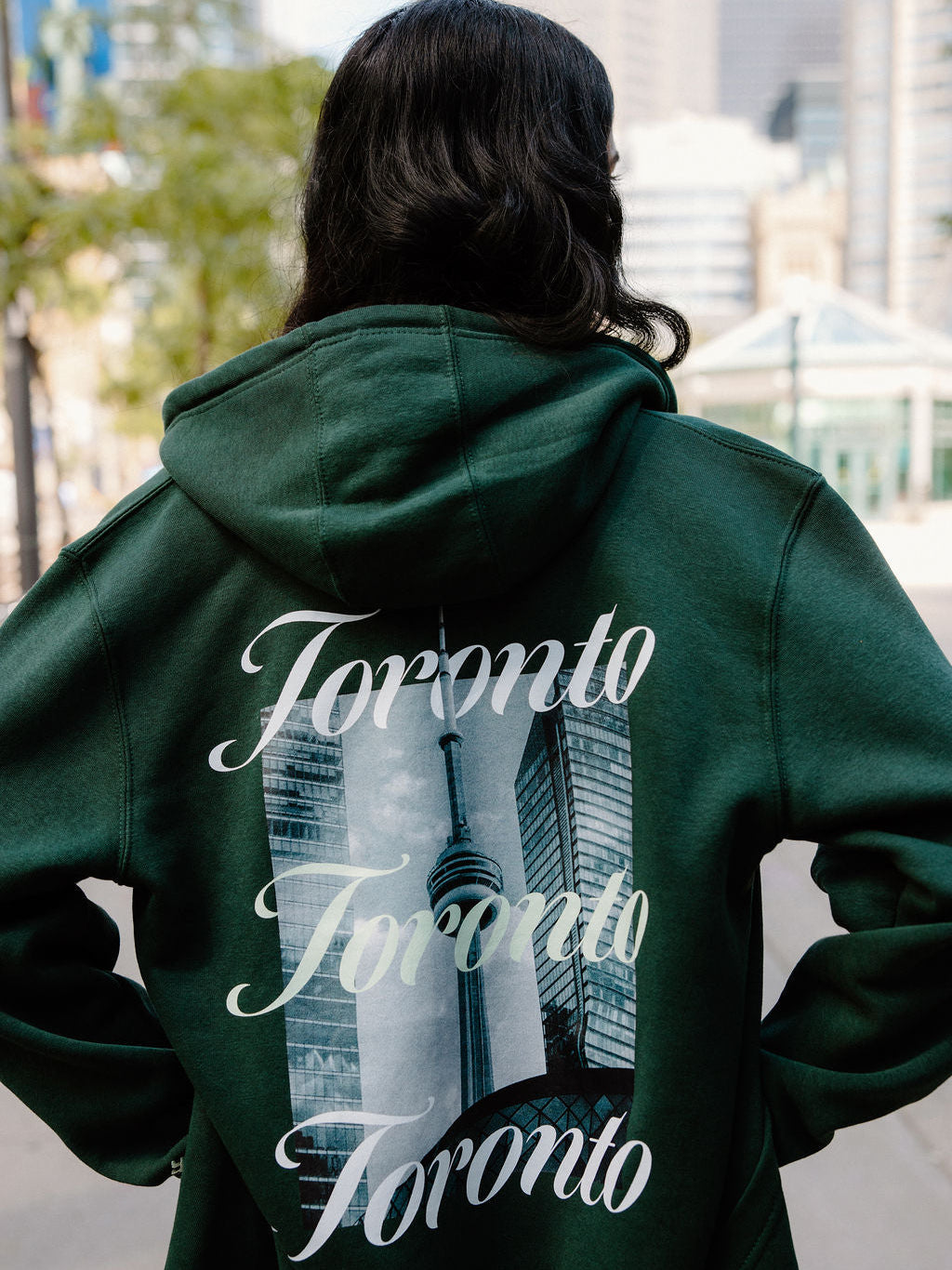 A person viewed from behind wearing a dark green hoodie with a large back graphic featuring the word 'Toronto' repeated in white script over a city skyline and the CN Tower.