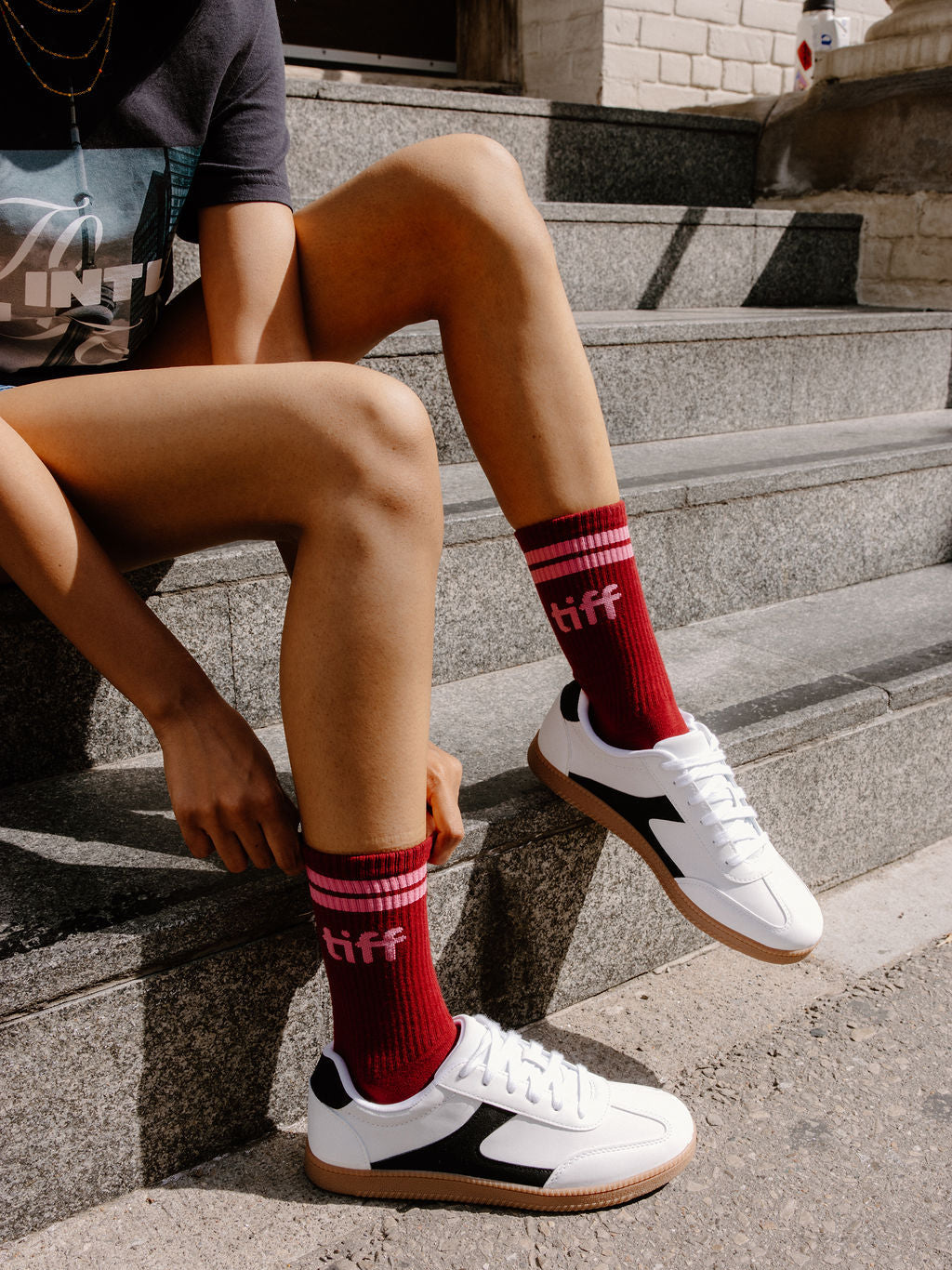 A person sitting on outdoor stone steps wearing red crew socks with white and black low-top sneakers. The socks have pink stripes at the top and the word 'tiff' printed in white near the cuff.