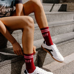 A person sitting on outdoor stone steps wearing red crew socks with white and black low-top sneakers. The socks have pink stripes at the top and the word 'tiff' printed in white near the cuff.