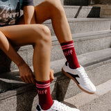 A person sitting on outdoor stone steps wearing red crew socks with white and black low-top sneakers. The socks have pink stripes at the top and the word 'tiff' printed in white near the cuff.
