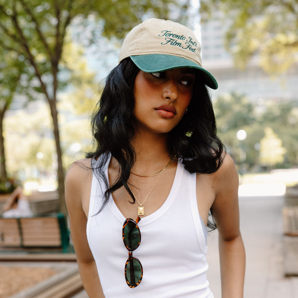 A woman wearing a beige and green baseball cap with 'Toronto Film Festival' embroidered on it, a white ribbed tank top, and white jeans, standing outdoors in a park-like setting.
