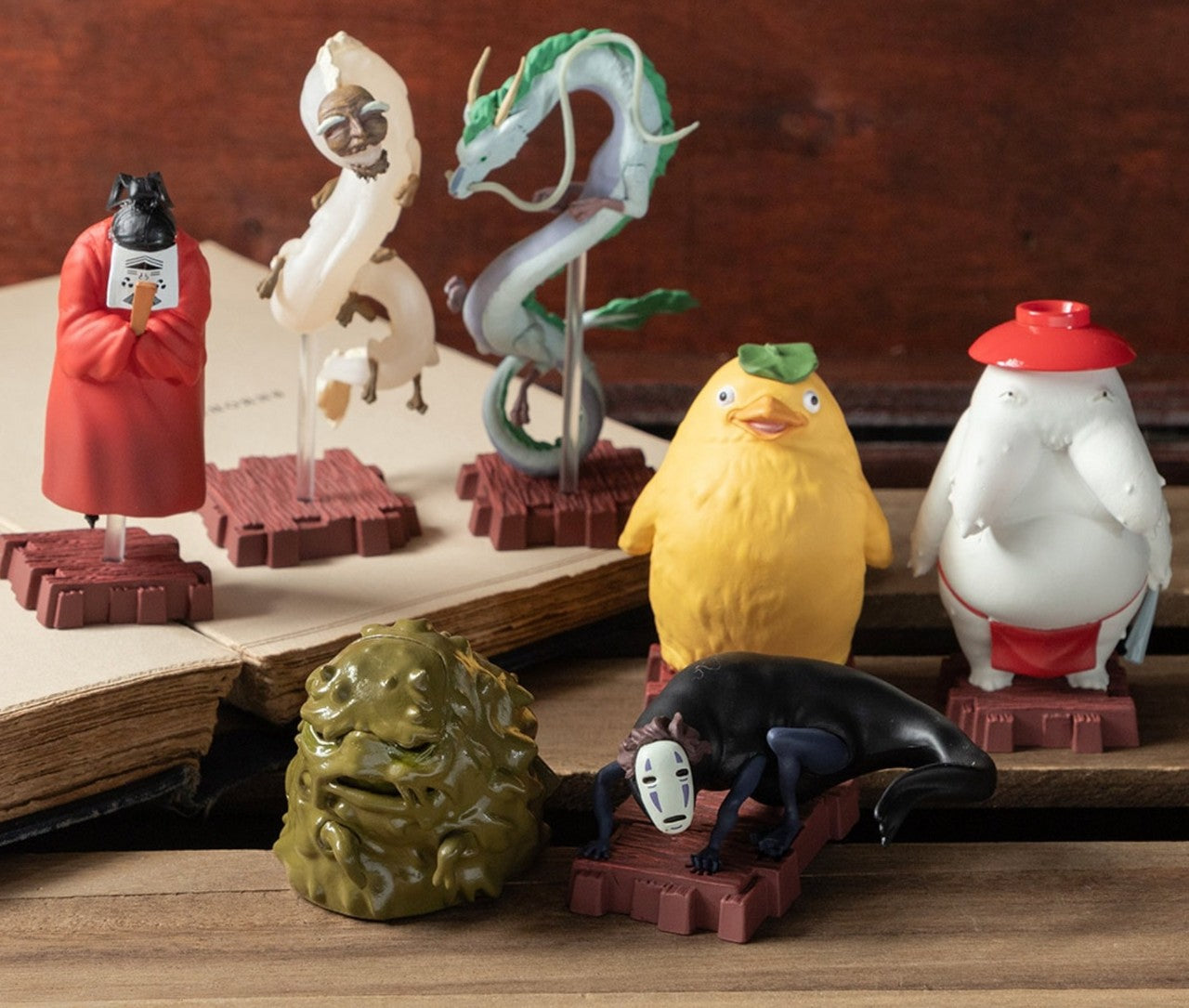 Small figurines depicting various spirits and creatures from the Studio Ghibli film Spirited Away - all arranged on a wooden surface around an open book.