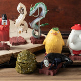 Small figurines depicting various spirits and creatures from the Studio Ghibli film Spirited Away - all arranged on a wooden surface around an open book.