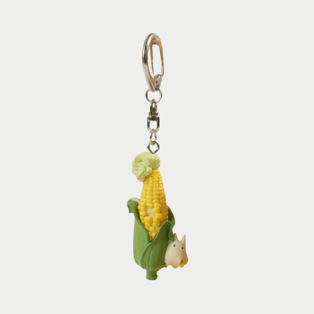 A figural keychain featuring a yellow cob of corn wrapped in green husks, with a tiny white Totoro peeking out from the bottom of the leaves, all suspended from a silver metal clip.