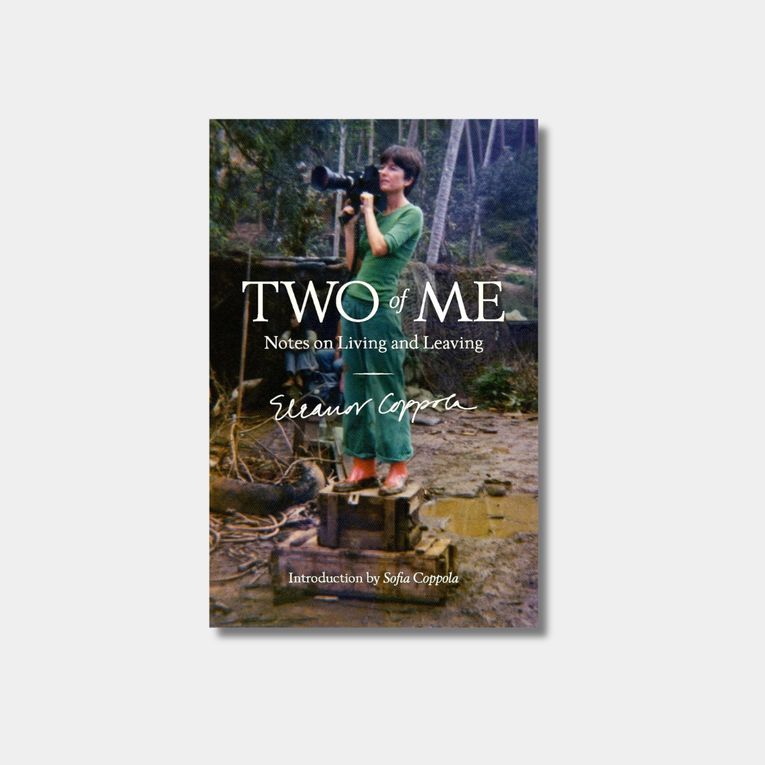The cover of the book "Two of Me: Notes on Living and Leaving" features a vintage photo of Eleanor Coppola standing on a film set in a long green dress, holding a large movie camera.