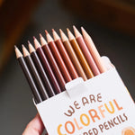 A hand holds an open box of "We Are Colorful" colored pencils, revealing twelve sharpened tips in a range of diverse skin tones.