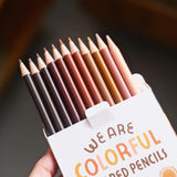A hand holds an open box of "We Are Colorful" colored pencils, revealing twelve sharpened tips in a range of diverse skin tones.