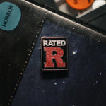 A rectangular enamel pin mimicking a retro "Rated R" movie rating box, featuring a large, textured red letter "R" beneath the white word "RATED" on a black background, sits pinned onto a dark surface near a blue "HORROR" genre sticker.