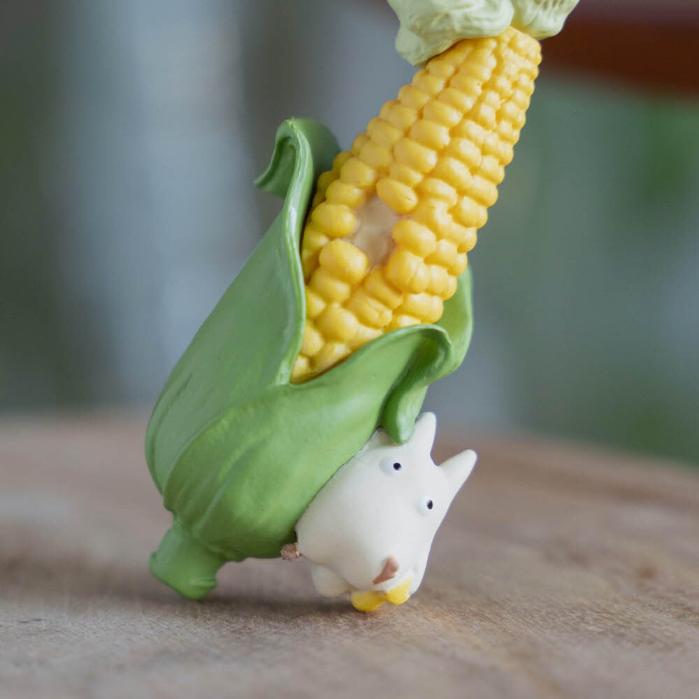 A close-up image showing two sides of a figural corn cob keychain: one side features a tiny white Totoro peeking from the bottom of the green husk, while the other side reveals a small, round black Soot Sprite tucked among the yellow corn kernels.