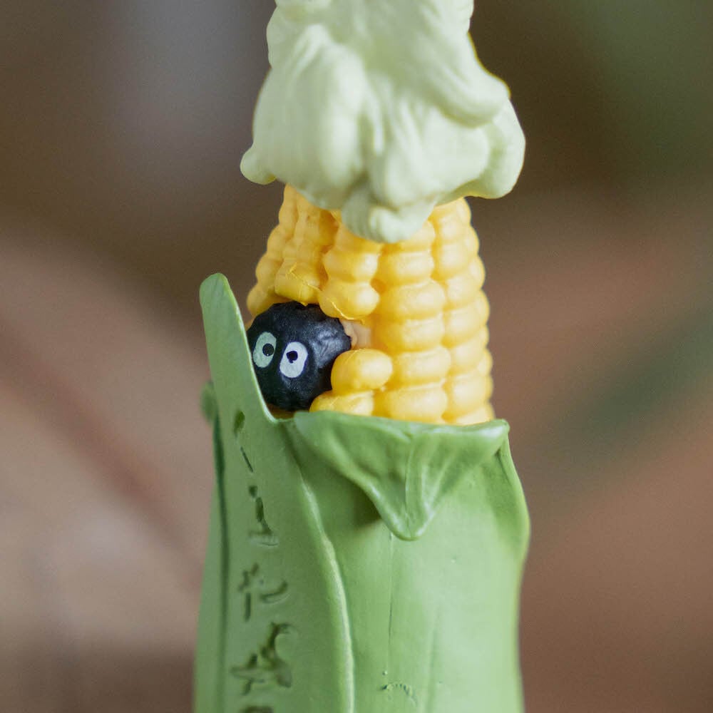 A close-up image showing two sides of a figural corn cob keychain: one side features a tiny white Totoro peeking from the bottom of the green husk, while the other side reveals a small, round black Soot Sprite tucked among the yellow corn kernels.