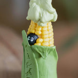 A close-up image showing two sides of a figural corn cob keychain: one side features a tiny white Totoro peeking from the bottom of the green husk, while the other side reveals a small, round black Soot Sprite tucked among the yellow corn kernels.