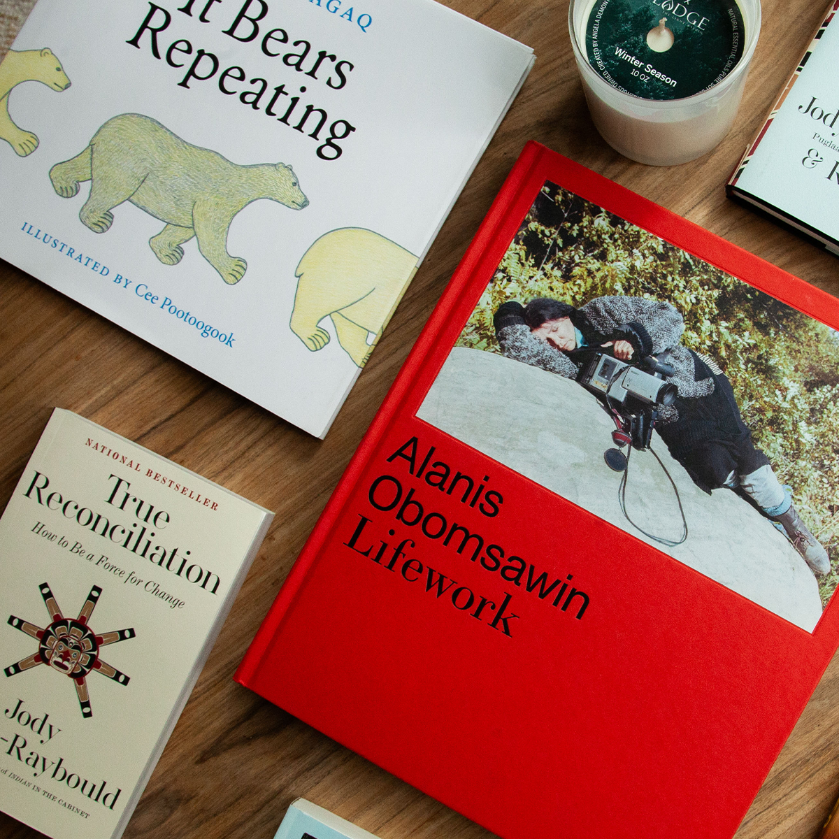 An assortment of books by Indigenous authors and illustrators, including titles on reconciliation and filmmaker Alanis Obomsawin, are arranged on a wooden surface next to a candle.