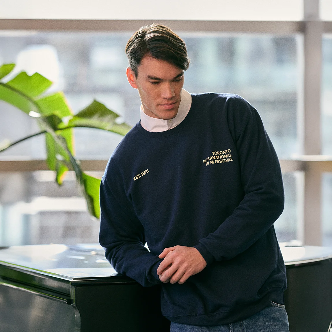 Man wearing a navy blue sweatshirt with text, standing indoors with a plant in the background