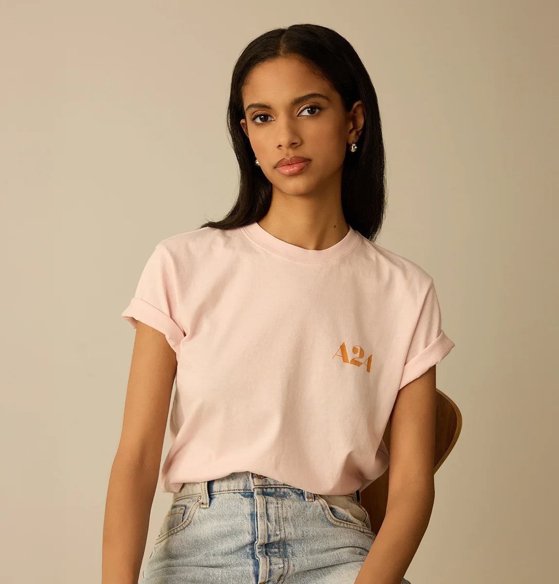 Woman wearing a light pink t-shirt with a logo, sitting against a beige background