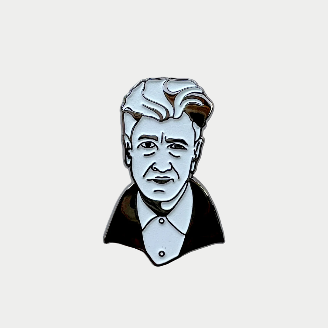 An enamel lapel pin depicting a monochrome bust illustration of filmmaker David Lynch with his signature swept-back white hair, a dark jacket, and a buttoned shirt against a plain gray background.