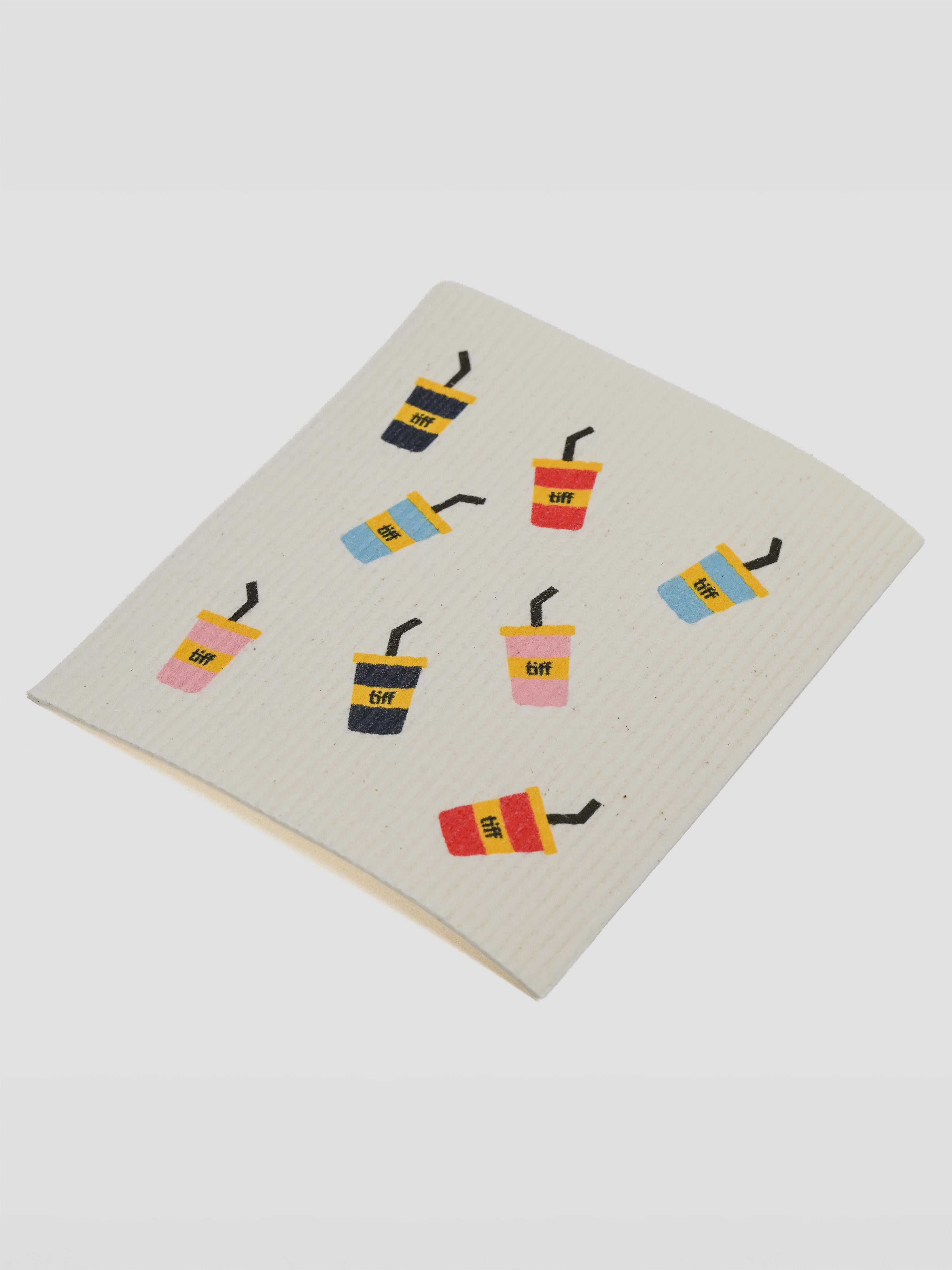 A square cream-colored dishcloth with a waffle texture, printed with a pattern of colorful iced coffee cups, on a light gray background.
