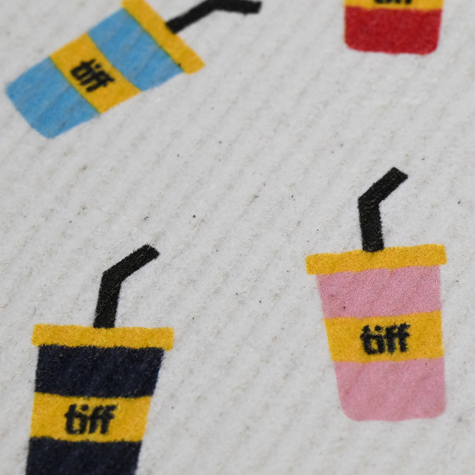A close-up of a textured white fabric featuring a repeating pattern of colorful drink cups with yellow bands and the text 'tiff' printed in black.