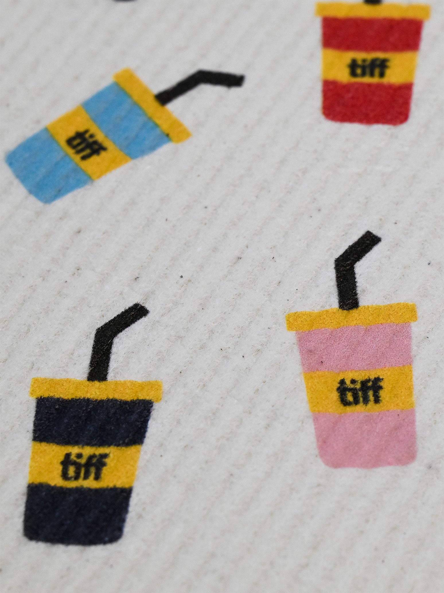 A close-up of a textured white fabric featuring a repeating pattern of colorful drink cups with yellow bands and the text 'tiff' printed in black.