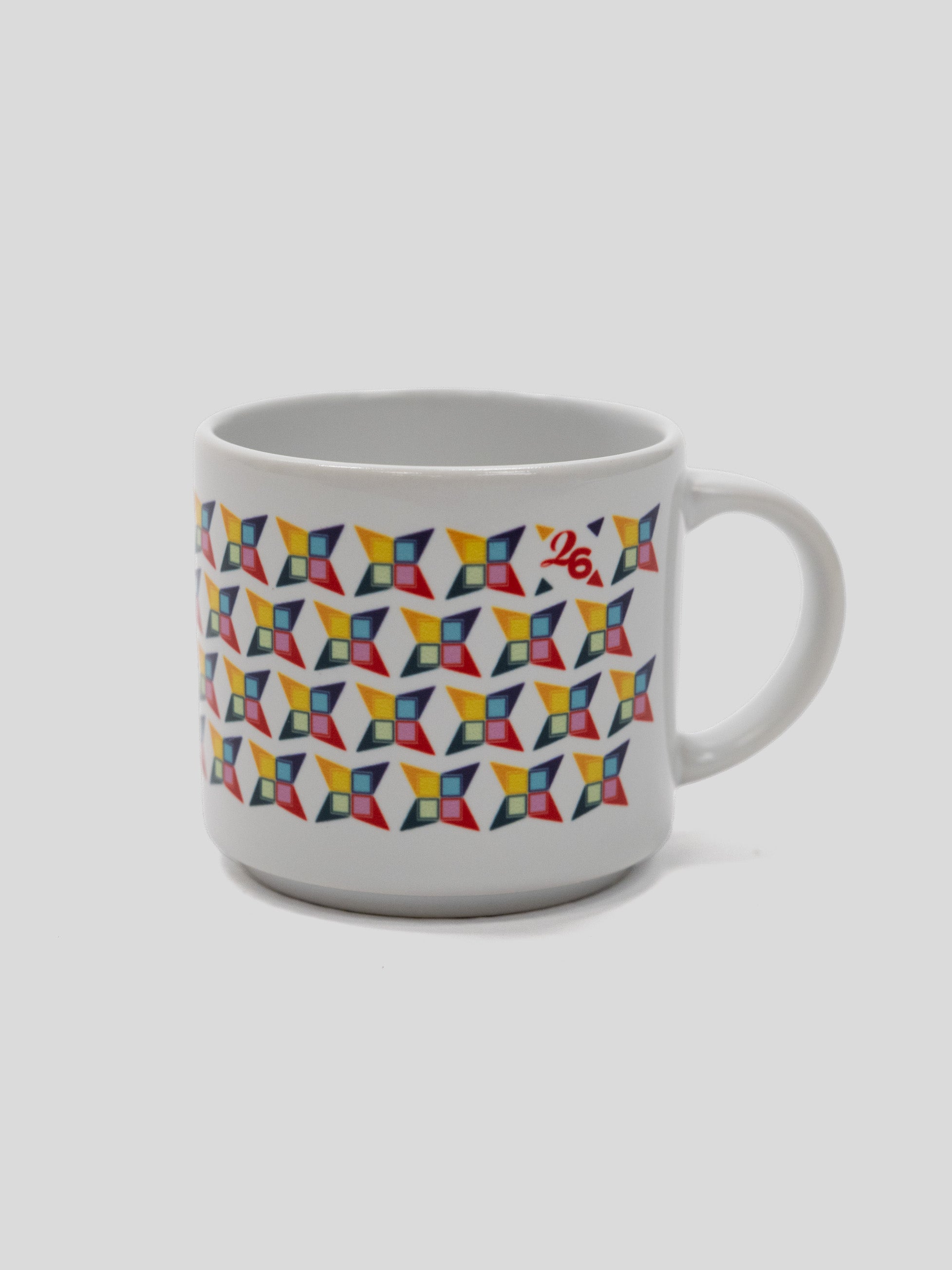 A white ceramic coffee mug featuring a colorful geometric pattern of repeating 3D cube-like shapes in red, yellow, blue, and green, with a small red logo on the upper right side, set against a solid light gray background.
