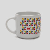 A white ceramic coffee mug featuring a repeating geometric pattern of colorful pinwheel-like shapes in yellow, orange, red, blue, and black, with a small red logo in the center.