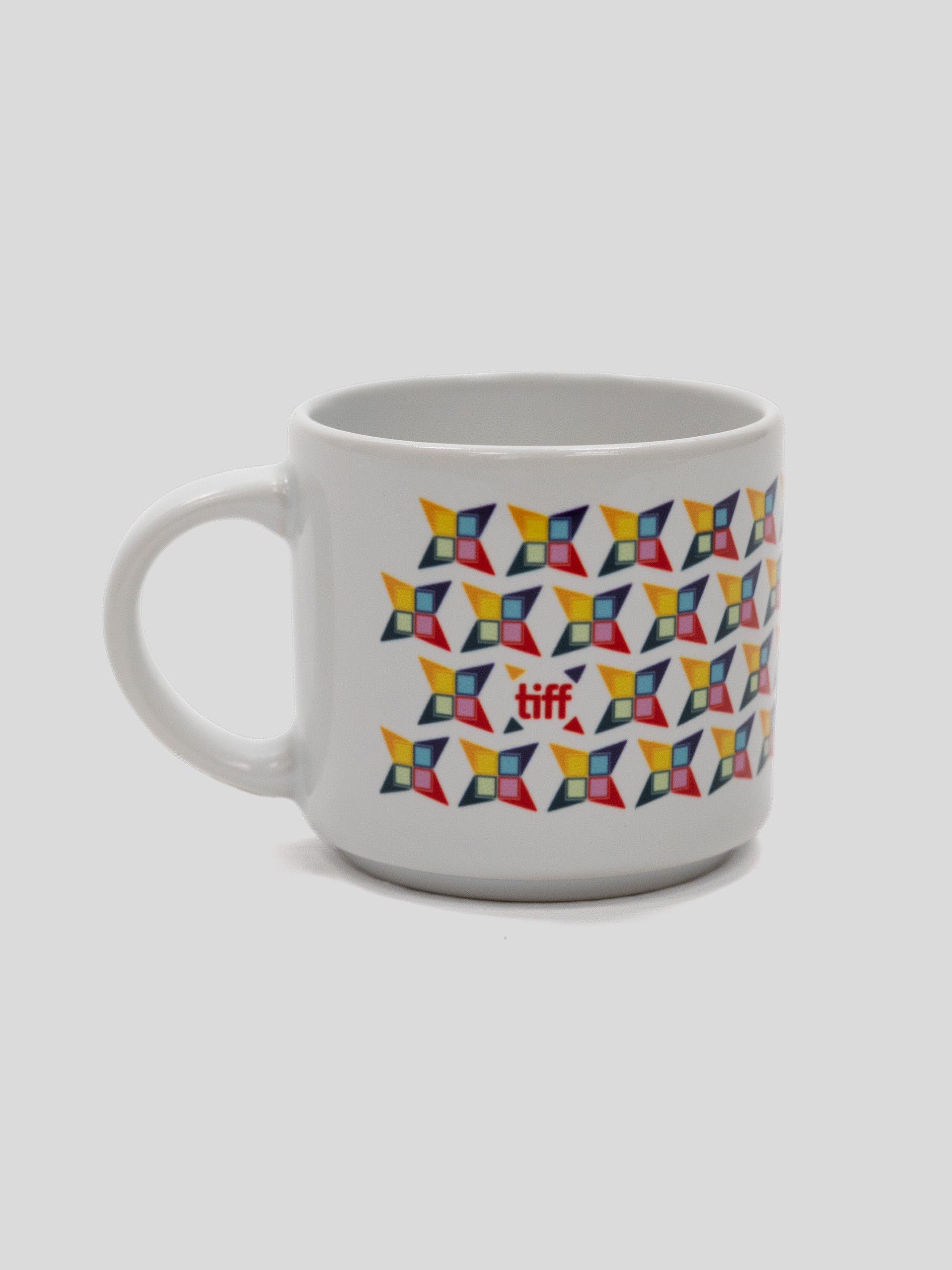 A white ceramic coffee mug featuring a repeating geometric pattern of colorful pinwheel-like shapes in yellow, orange, red, blue, and black, with a small red logo in the center.