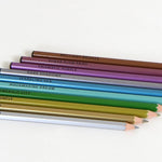 A row of ten sharpened metallic colored pencils arranged diagonally, showcasing a range of shimmering tones from silver and gold to emerald green and deep plum.