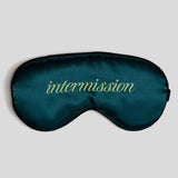A dark teal satin sleep mask with the word 'intermission' embroidered in a light yellow-green cursive font across the center, shown against a solid light gray background.