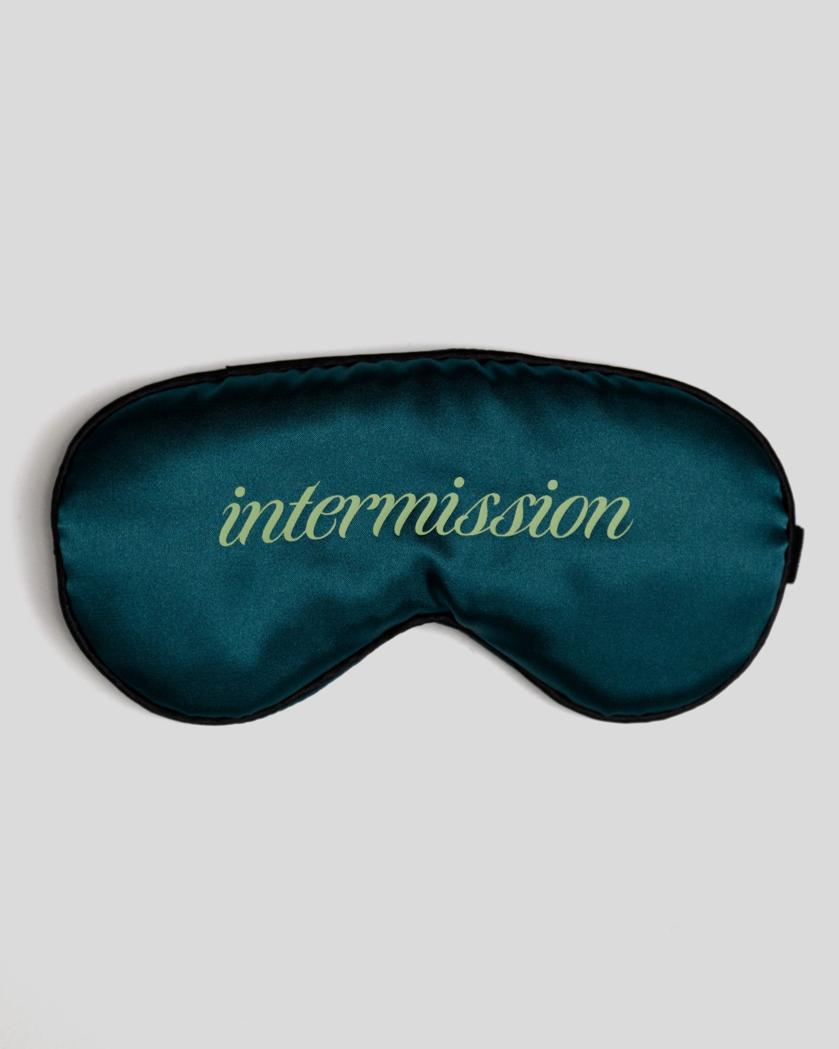 A dark teal satin sleep mask with the word 'intermission' embroidered in a light yellow-green cursive font across the center, shown against a solid light gray background.