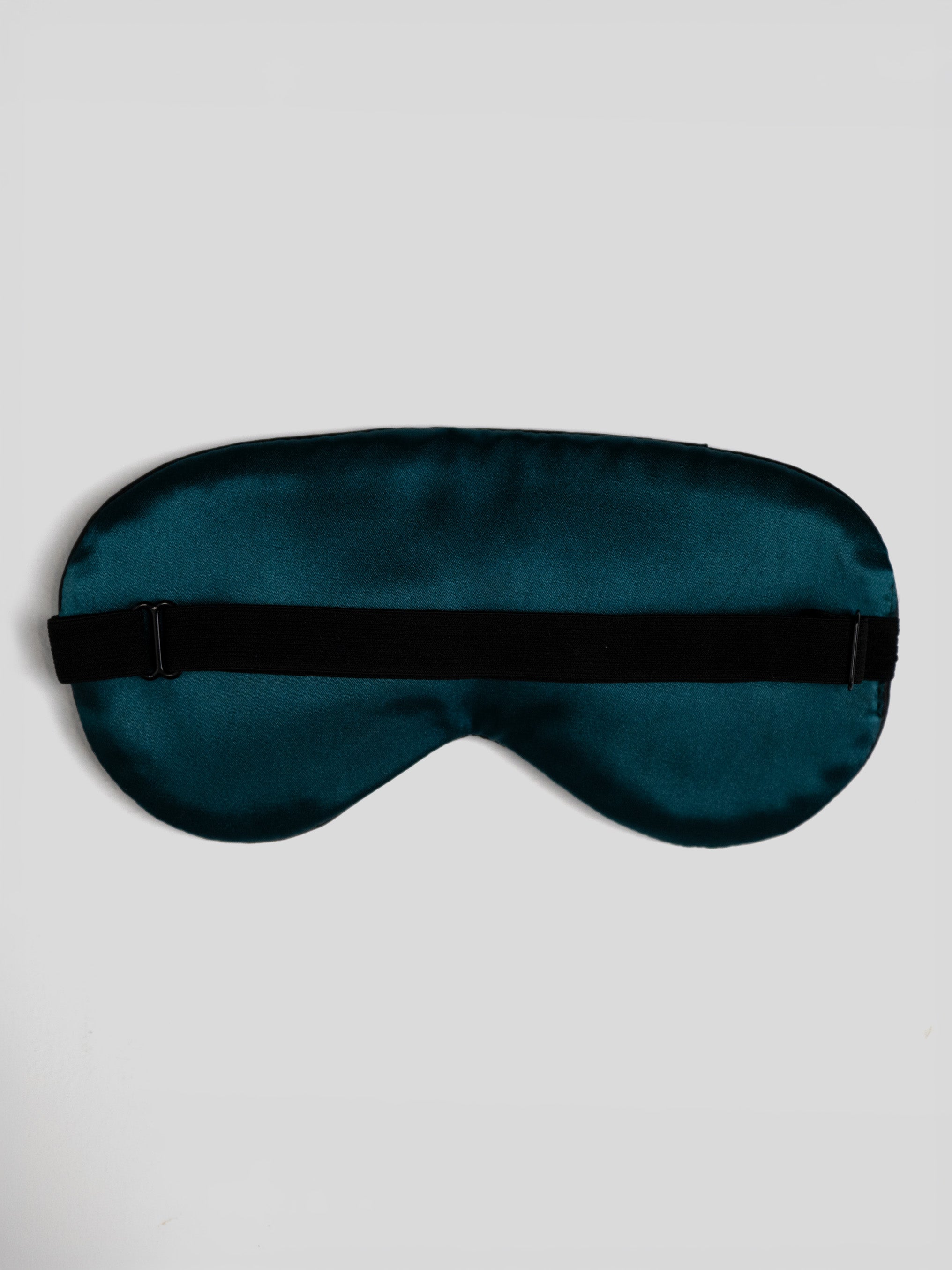 A dark teal satin sleep mask with a black elastic strap, laid flat against a light gray background.