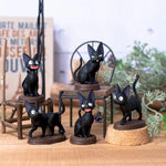 Five miniature figurines of the black cat Jiji from Kiki's Delivery Service are arranged on a wooden surface around a tiny metal chair and wooden stool, surrounded by soft-focus greenery and signs.