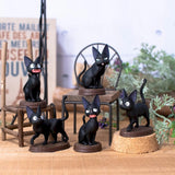 Five miniature figurines of the black cat Jiji from Kiki's Delivery Service are arranged on a wooden surface around a tiny metal chair and wooden stool, surrounded by soft-focus greenery and signs.
