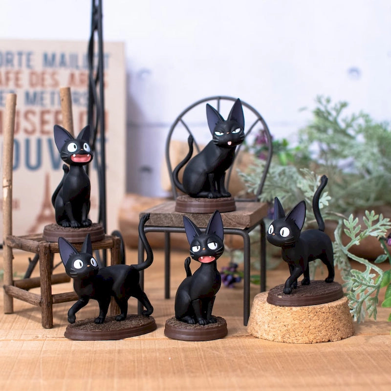 Five miniature figurines of the black cat Jiji from Kiki's Delivery Service are arranged on a wooden surface around a tiny metal chair and wooden stool, surrounded by soft-focus greenery and signs.