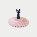 A black Jiji figurine stands in the center of a scalloped pink silicone mug lid.